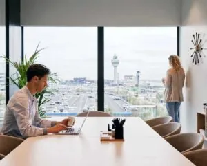 Man werkt op laptop aan vergadertafel terwijl vrouw uitkijkt op Schiphol vanachter kantoorraam.