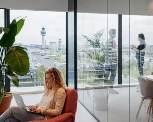Vrouw werkt op een laptop in een modern kantoor met uitzicht op de verkeerstoren van Schiphol.