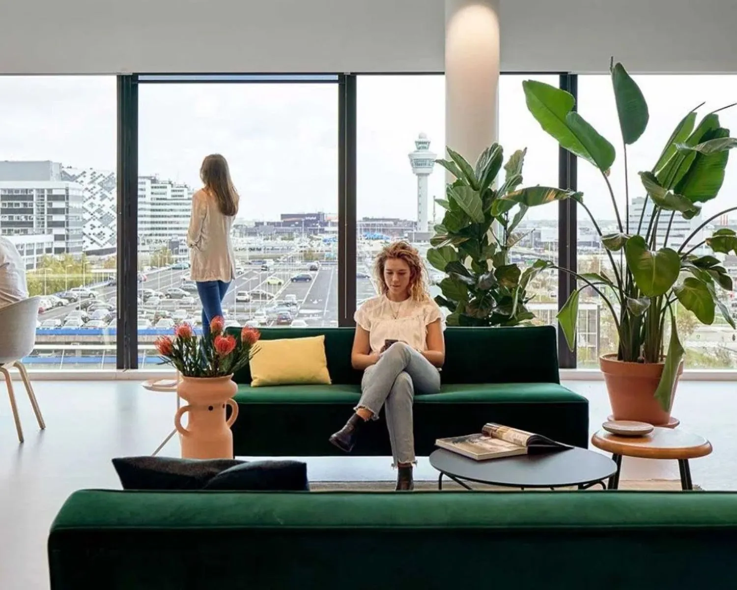 Moderne wachtruimte met uitzicht op Schiphol, waar een vrouw op een groene bank zit en een andere vrouw uit het raam kijkt.