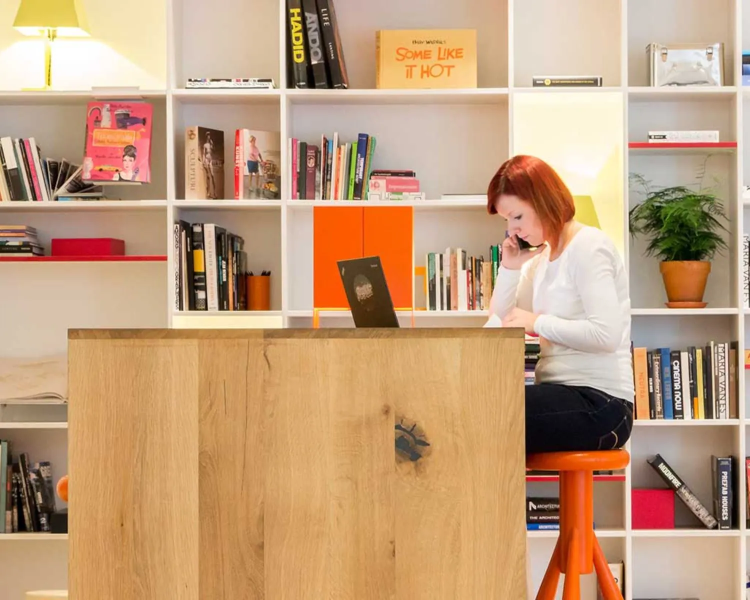 Vrouw met rood haar werkt aan een laptop en telefoneert in een moderne bibliotheek met boekenplanken op de achtergrond.