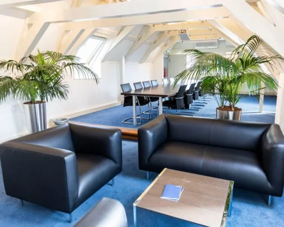 Moderne vergaderruimte met zwarte leren zitbanken, een lange vergadertafel, kamerplanten en een blauw tapijt.