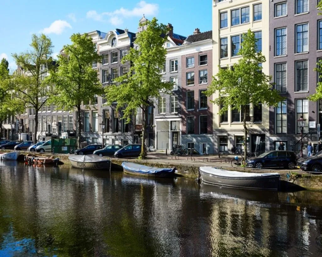 Woonboten en geparkeerde auto's langs een gracht met hoge herenhuizen in Amsterdam.
