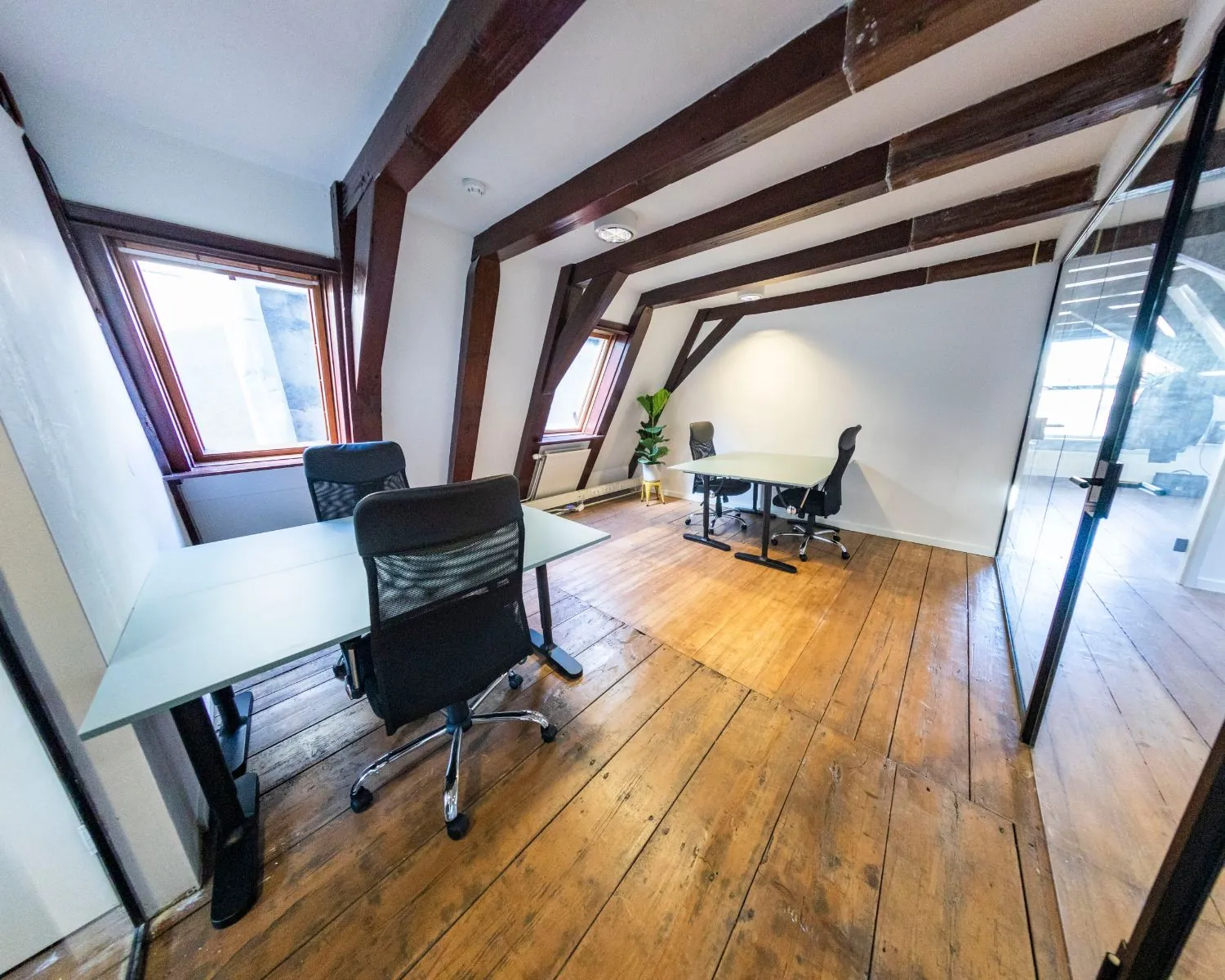 Kantoorruimte met houten vloer, schuine balken, bureaus en bureaustoelen in een lichte zolderkamer.