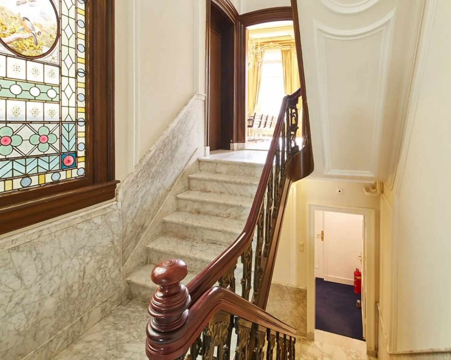 Marmeren trappenhuis met houten leuning, glas-in-loodraam en zicht op een zonnige kamer bovenaan.