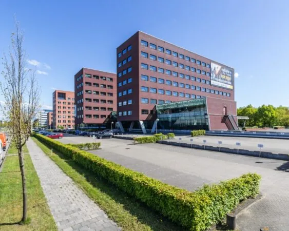 Modern kantoorgebouw met rode bakstenen en glazen entree, omringd door parkeerplaatsen en groenvoorziening.