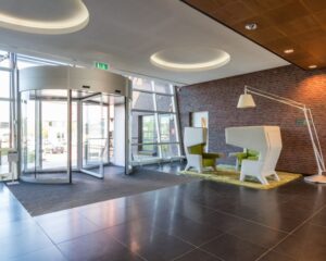 Moderne entreehal met draaideur, designstoelen en een grote vloerlamp tegen een bakstenen muur.