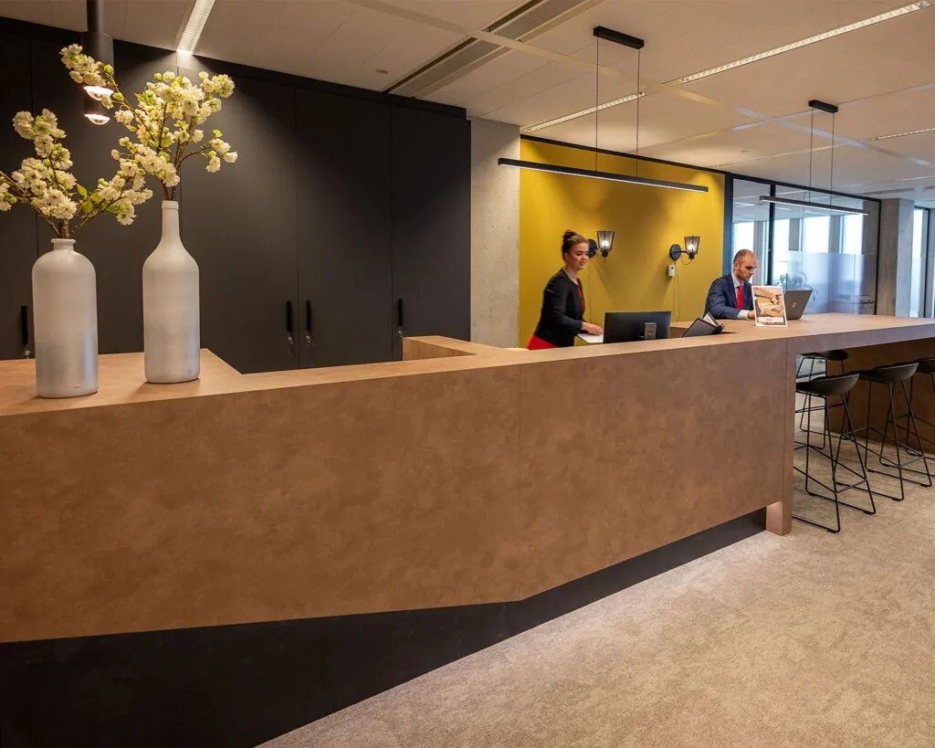 Moderne receptiebalie met twee medewerkers, gele accentmuur en decoratieve vazen met bloemen.
