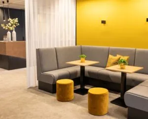 Moderne zithoek met grijze banken, gele accenten en twee houten tafeltjes met plantjes.