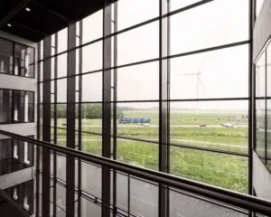Uitzicht vanuit een modern kantoorgebouw met glazen gevel op een groen landschap, wegverkeer en een windturbine.