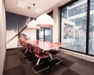 Vergaderruimte met een rode tafel, paarse stoelen, grote hanglampen en een muur met een windmolenafbeelding.