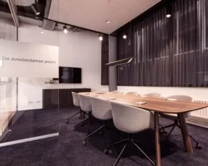 Vergaderruimte "De Amsterdamse poort" met een houten tafel, moderne stoelen en een flatscreen-tv aan de muur.