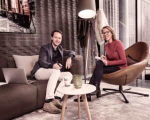 Twee mensen zitten in een moderne loungehoek en voeren een ontspannen gesprek met notitieboek en laptop.
