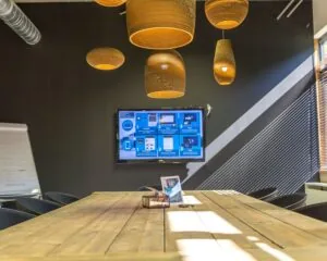 Vergaderruimte met houten tafel, hanglampen, een scherm met instructies en zonlicht dat door jaloezieën schijnt.