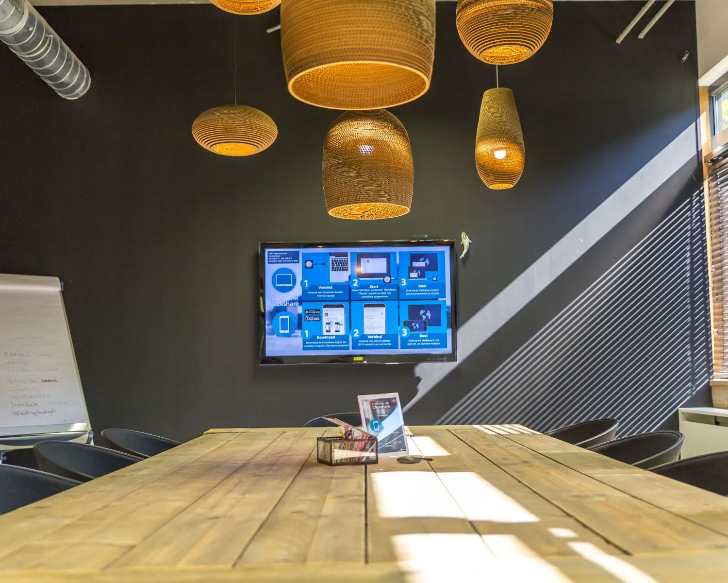 Vergaderruimte met houten tafel, hanglampen, een scherm met instructies en zonlicht dat door jaloezieën schijnt.