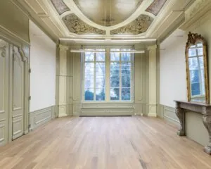 Elegante, klassieke kamer met hoog plafond, houten vloer, gedecoreerd plafond, grote ramen en een antieke spiegel boven een marmeren schouw.