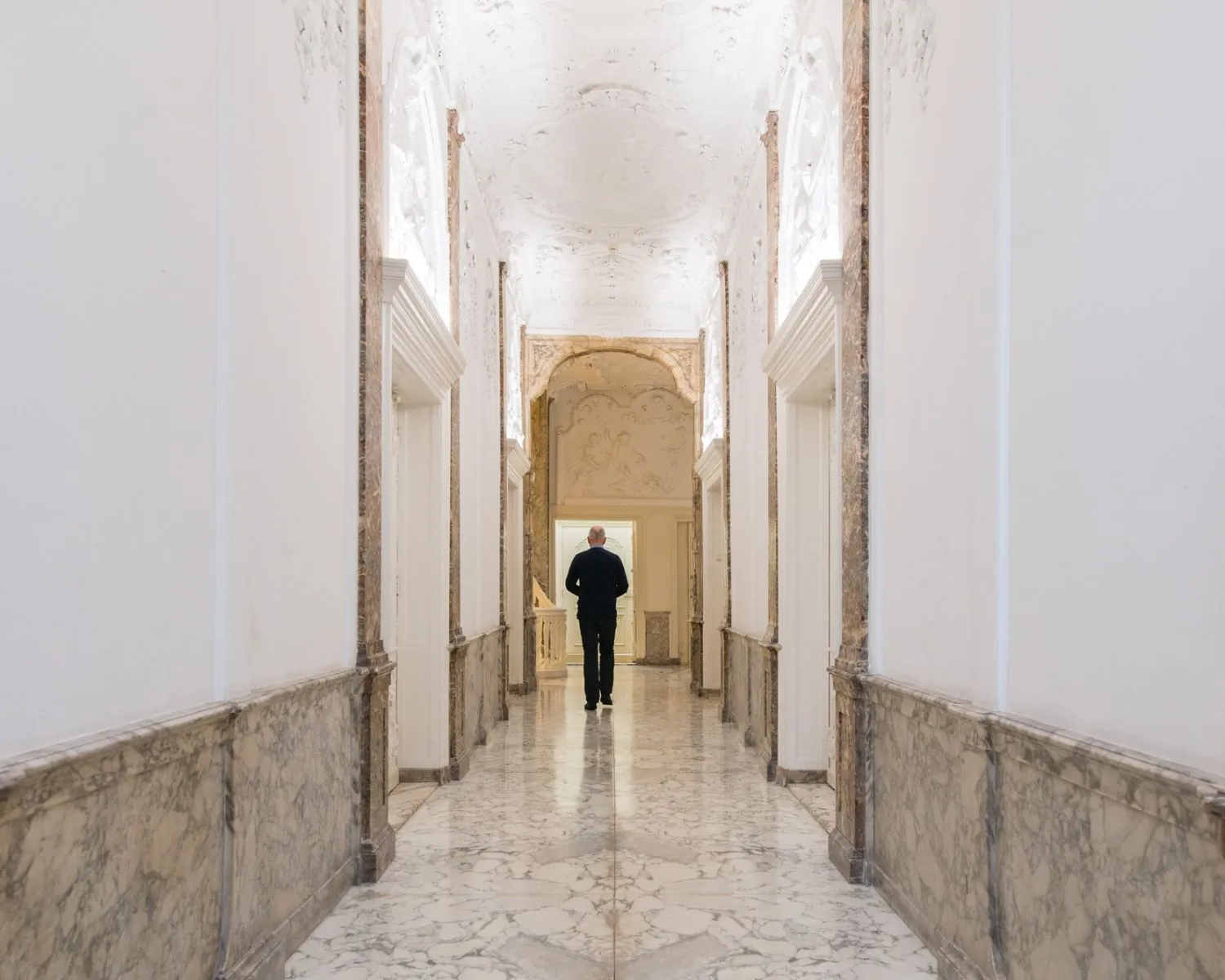 Man loopt door een marmeren gang met sierlijk wit plafond in een klassiek gebouw.