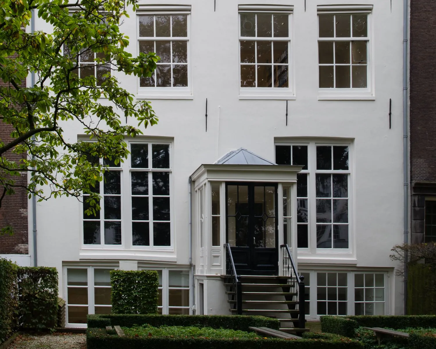 Achtergevel van een witgrachtenpand met symmetrische ramen, een veranda met trap en een formele tuin op de voorgrond.