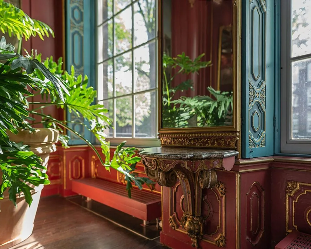 Een sierlijke, klassiek ingerichte kamer met grote planten, rood-blauw met gouden wanddecoratie en een spiegel boven een marmeren console.