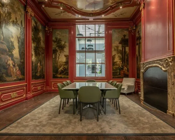 Vergaderruimte in klassieke stijl met rode wandpanelen, barokke schilderingen, een glazen tafel en moderne groene stoelen.