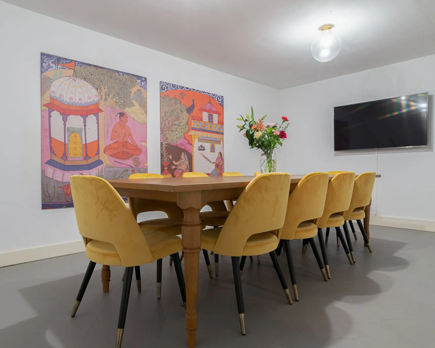 Vergaderruimte met een lange houten tafel, gele stoelen, kleurrijke Indiase kunst aan de muur en een flatscreen-tv.