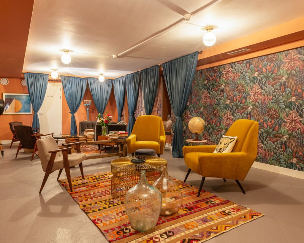 Stijlvol ingerichte lounge met kleurrijk vloerkleed, gele retrostoelen, glazen vazen en een botanisch behang.