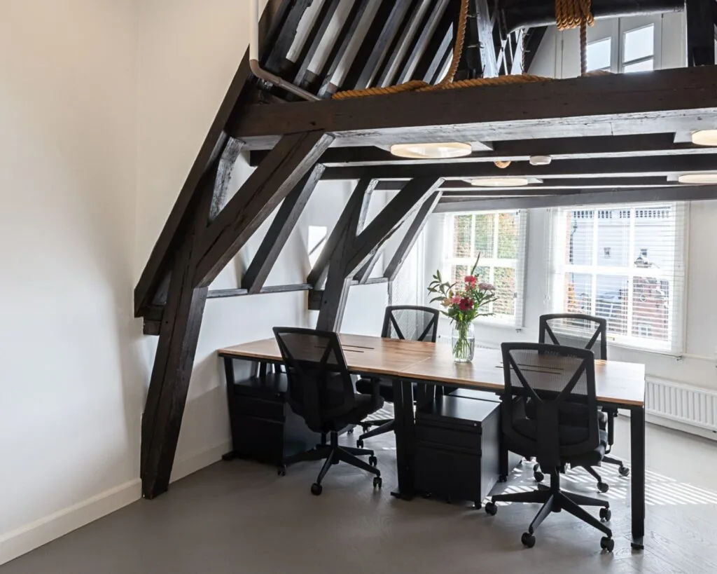 Moderne kantoorruimte met houten balken, een grote werktafel, zwarte bureaustoelen en een vaas bloemen bij het raam.