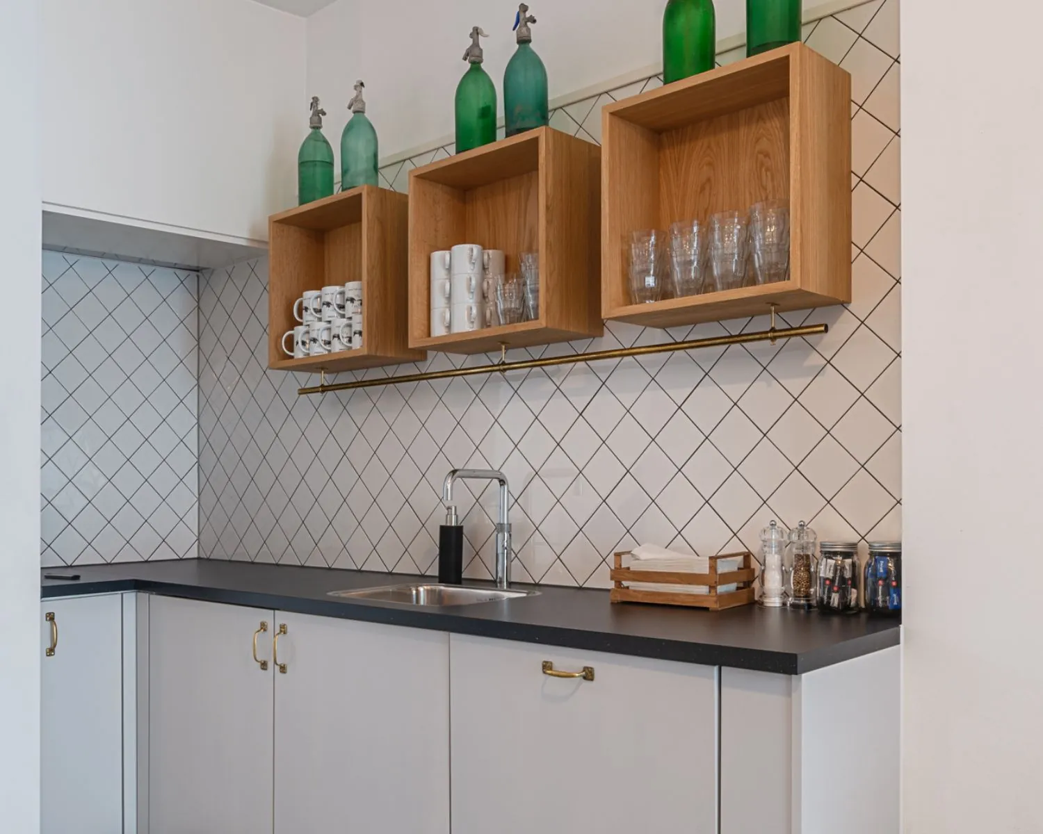 Moderne kitchenette met witte kastjes, een zwarte werkblad, houten wandrekken en groene vintage soda sifons als decoratie.