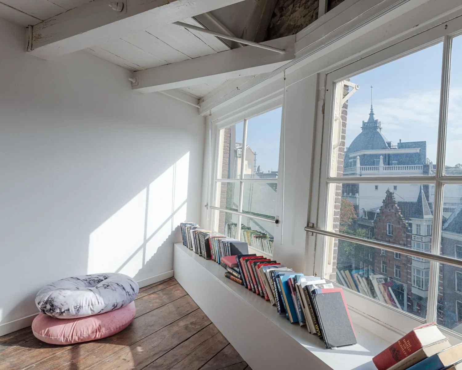 Lichte leeshoek met kussens, houten vloer en een vensterbank vol boeken met uitzicht op Amsterdamse gevels.