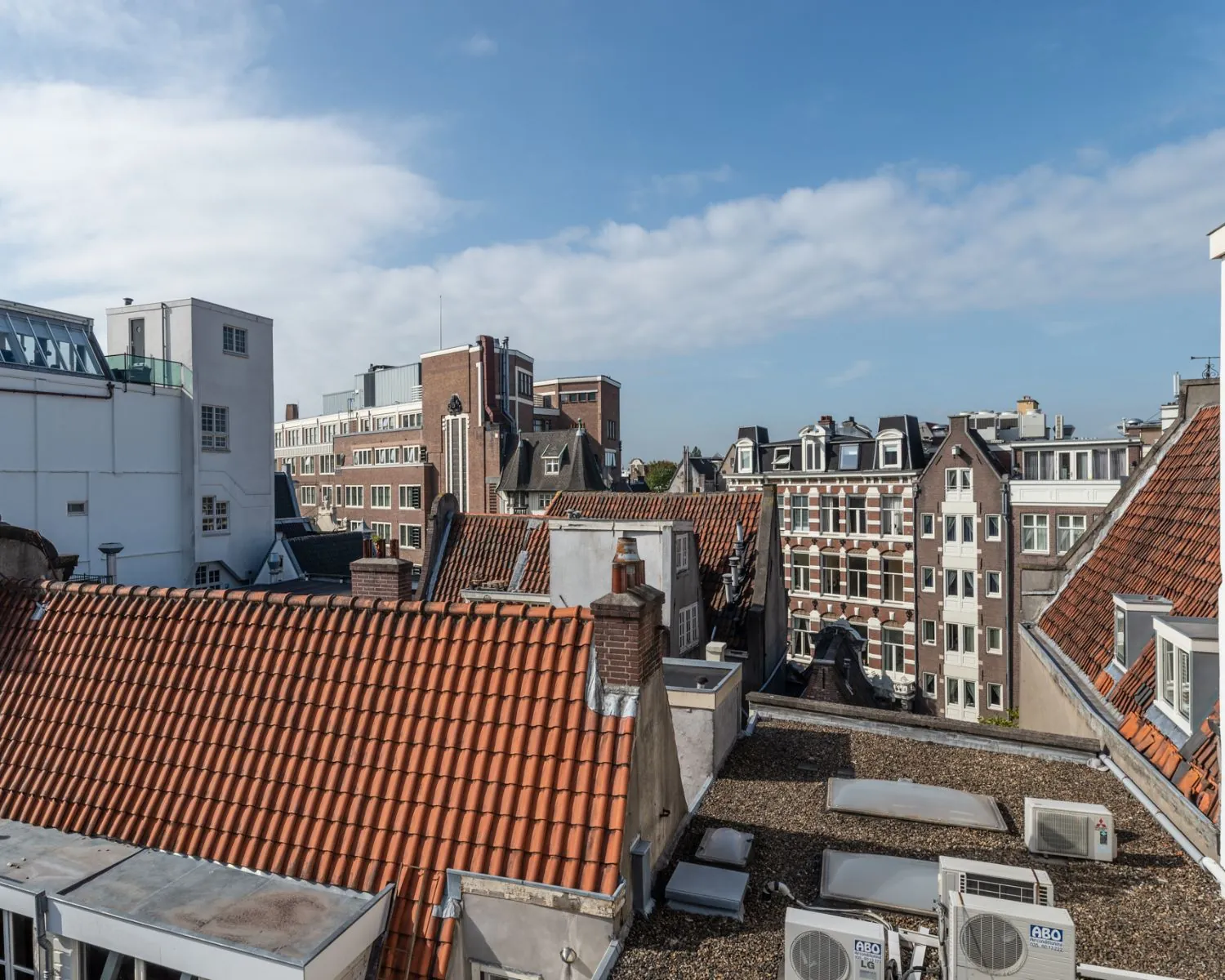 Uitzicht op Amsterdamse daken met rode dakpannen en achterliggende stadsgebouwen onder een deels bewolkte blauwe lucht.