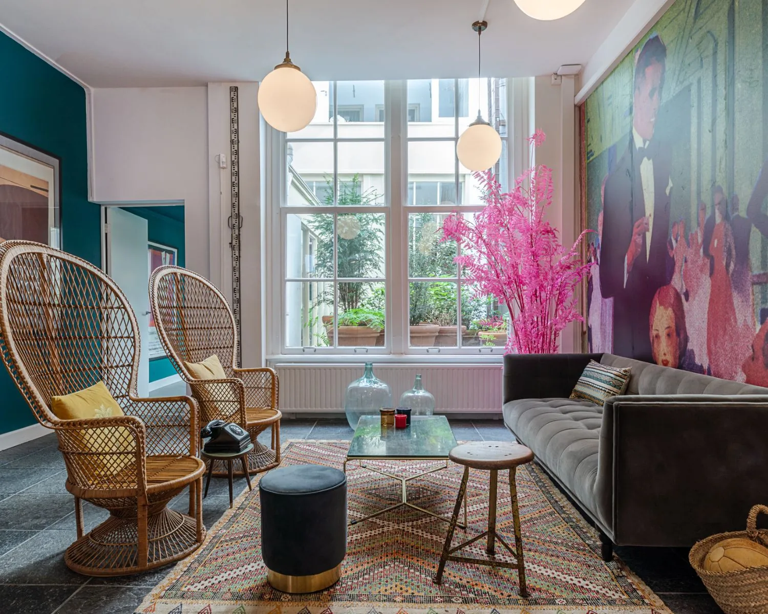 Stijlvol ingerichte woonkamer met rotanstoelen, een grijze bank, kleurrijk vloerkleed en een opvallende roze kunstplant naast een grote muurschildering.