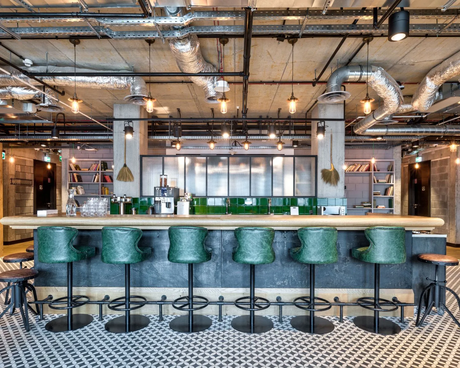 Moderne industriële barruimte met groene barkrukken, zichtbare leidingen en een betegelde vloer.