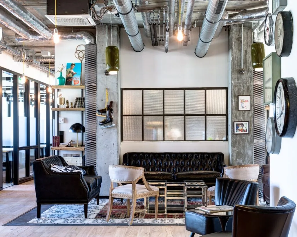 Industrieel ingerichte lounge met vintage meubels, decoratieve klokken en zichtbare ventilatiebuizen aan het plafond.