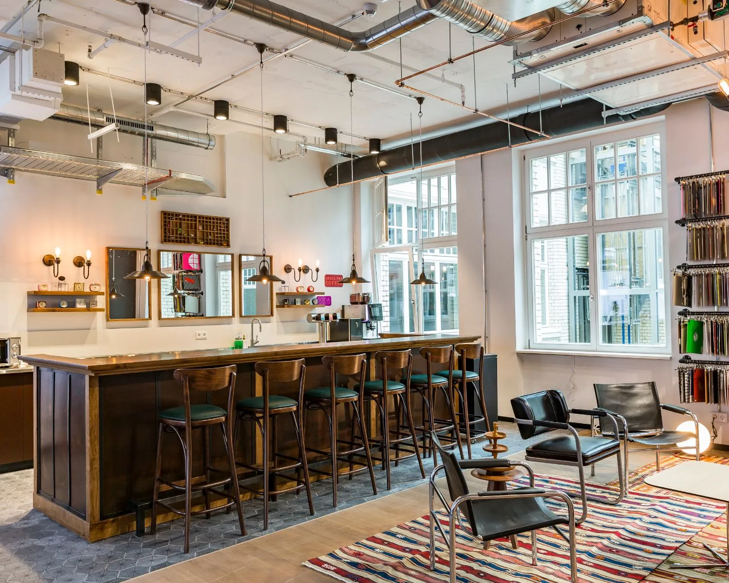 Modern ingerichte koffie- en ontmoetingsruimte met een houten bar, barkrukken, designstoelen en kleurrijk tapijt.