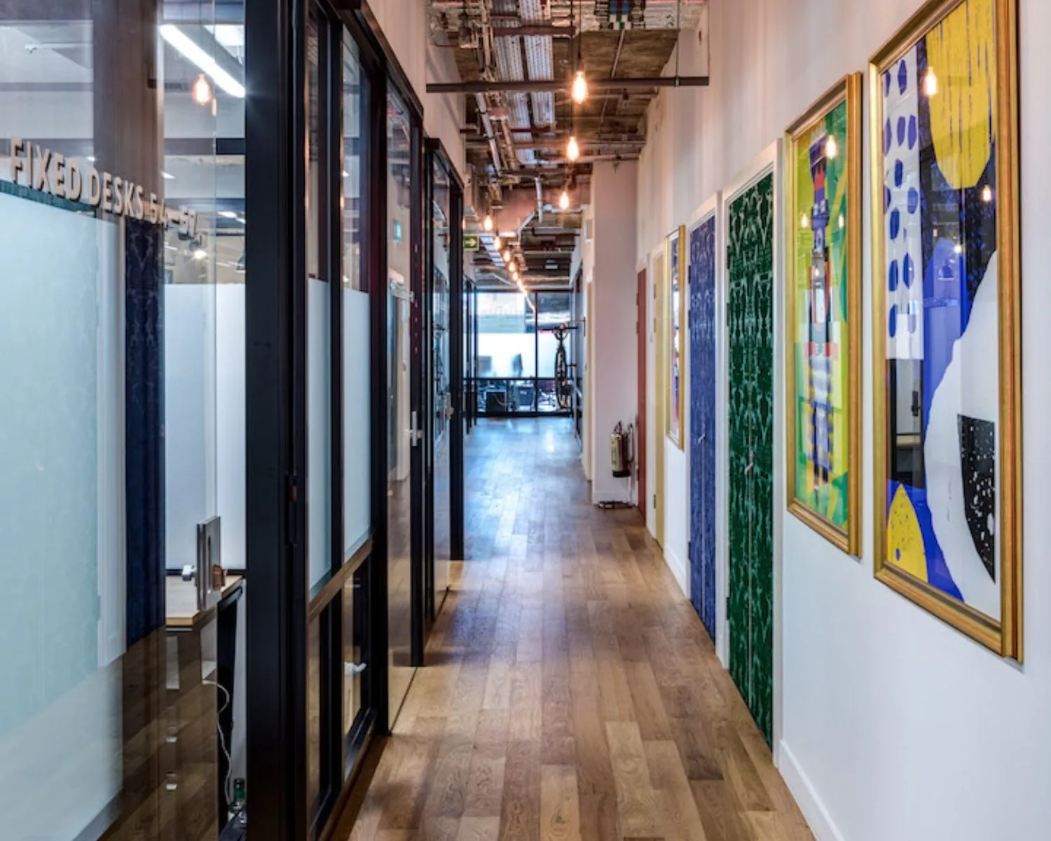 Gang in een modern kantoorpand met houten vloer, glazen kantoorwanden aan de linkerkant en kleurrijke kunstwerken aan de rechterwand.