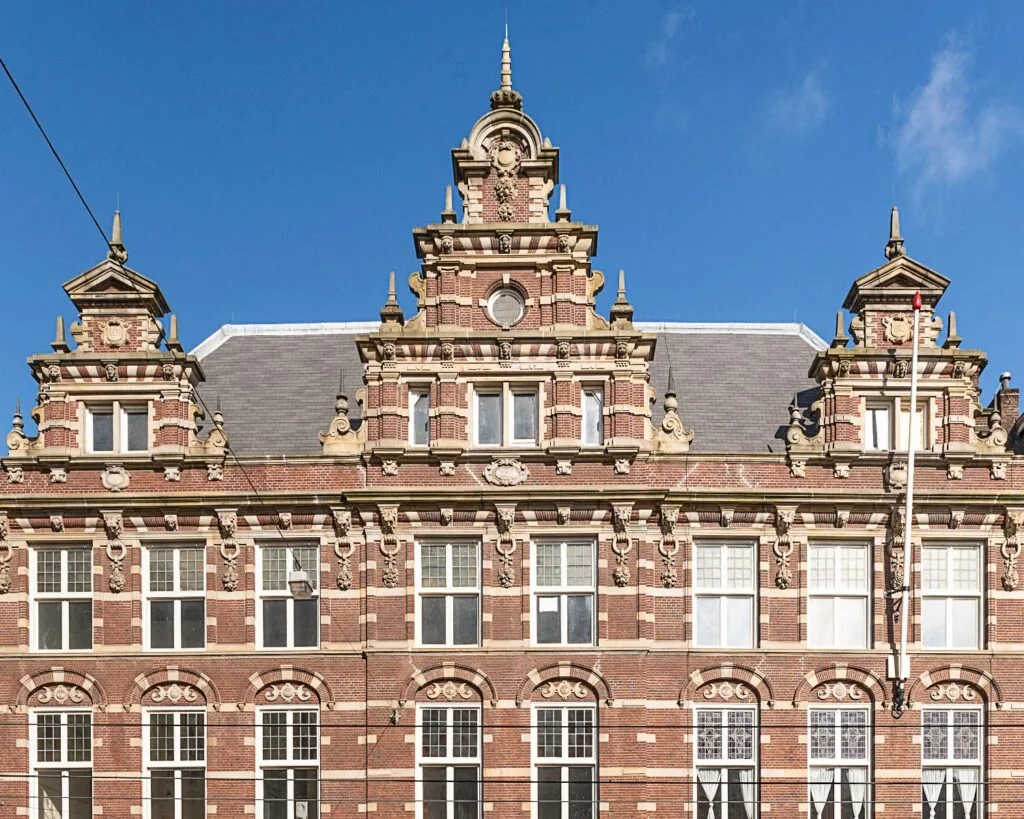 Voorgevel van een monumentaal, rijk versierd bakstenen pand met klassieke ornamenten en trapgevels tegen een blauwe lucht.