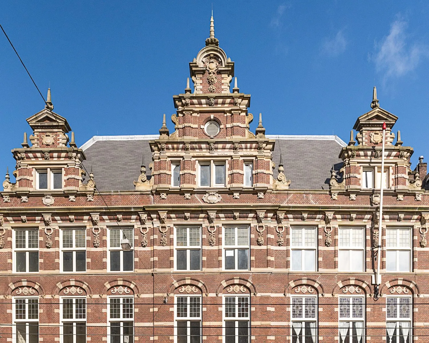 Voorgevel van een monumentaal, rijk versierd bakstenen pand met klassieke ornamenten en trapgevels tegen een blauwe lucht.