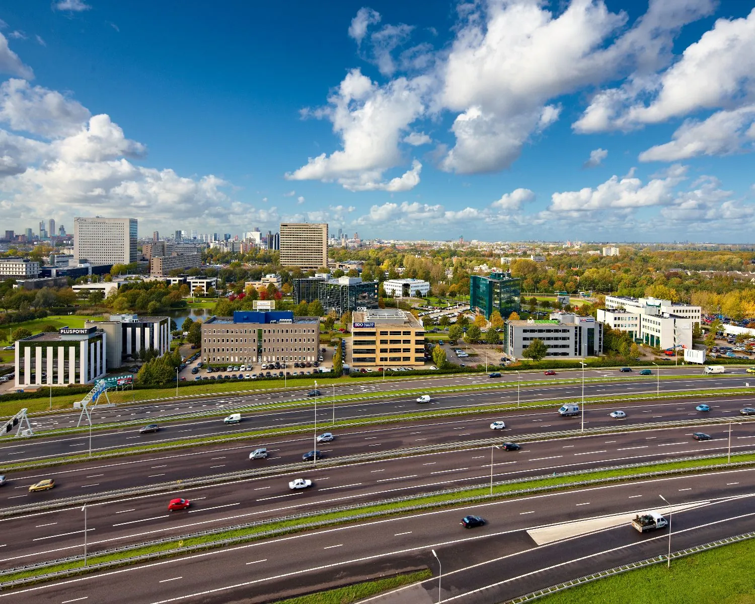 Luchtfoto van een snelweg met kantoorgebouwen in een stedelijk gebied onder een deels bewolkte blauwe lucht.