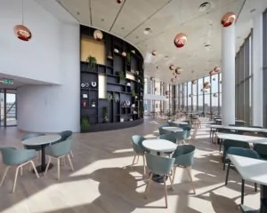 Moderne en lichtovergoten kantineruimte met hoge ramen, strakke meubels en een grote zwarte wand met planten en decoratie.