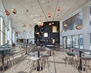 Moderne, lichtovergoten kantoorkantine met ronde tafels, houten barkrukken en opvallende hanglampen.