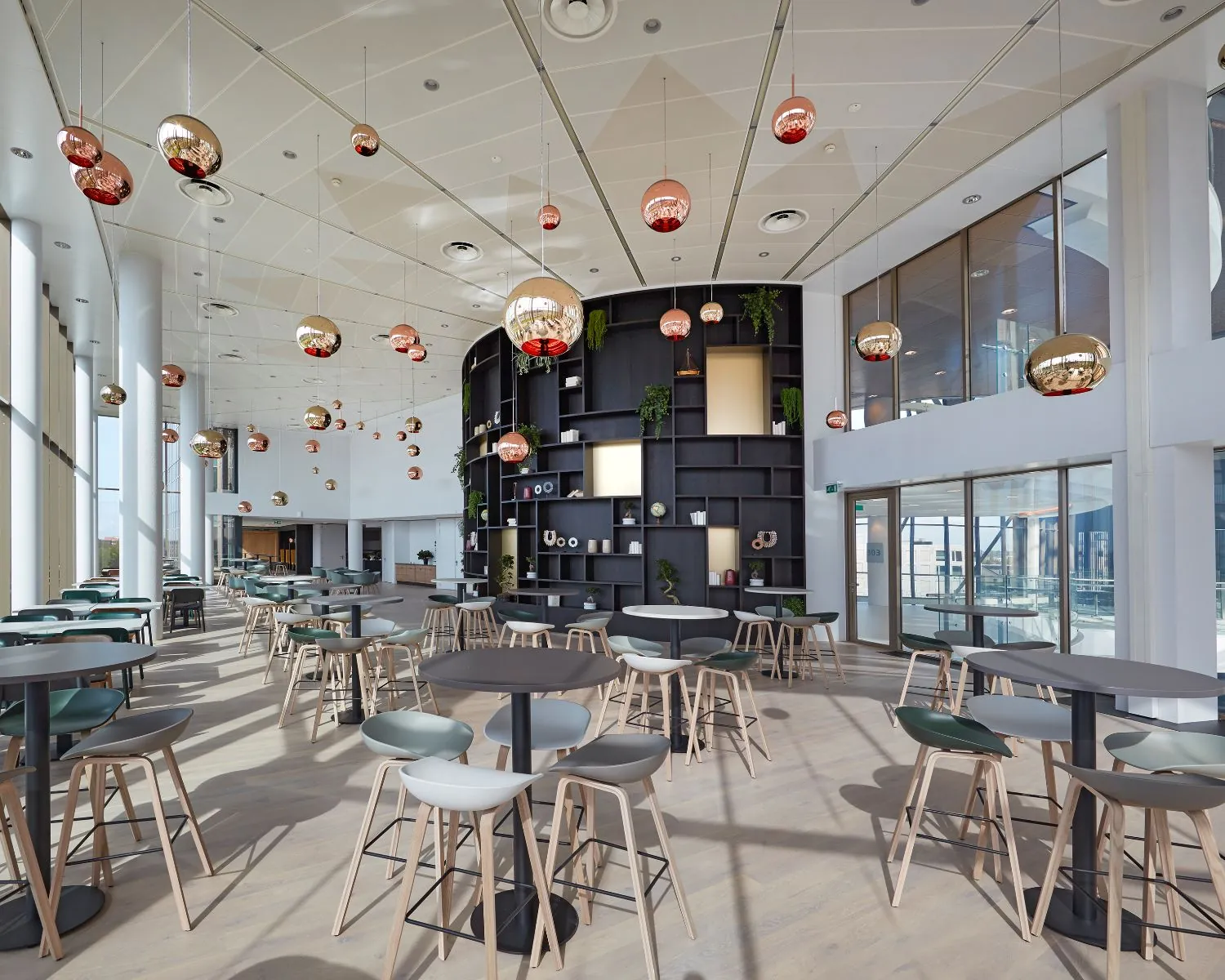 Moderne, lichtovergoten kantoorkantine met ronde tafels, houten barkrukken en opvallende hanglampen.