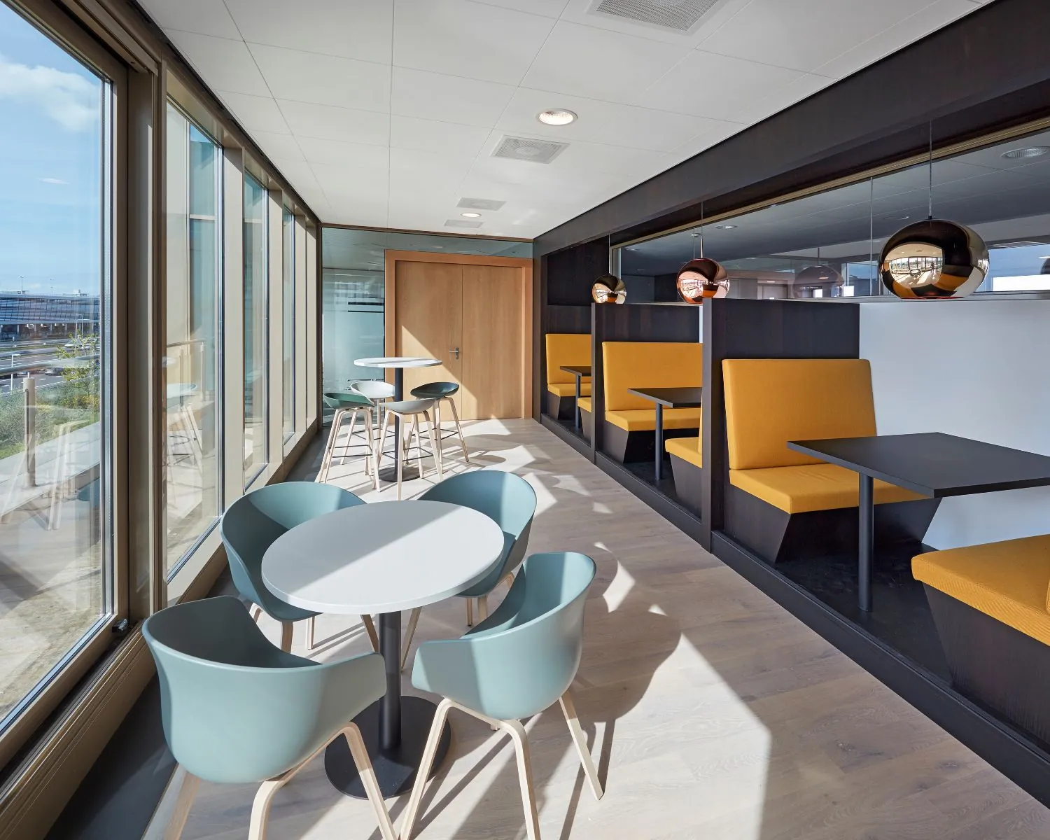 Moderne kantoorlounge met zitnissen, ronde tafels en grote ramen met uitzicht op buiten.