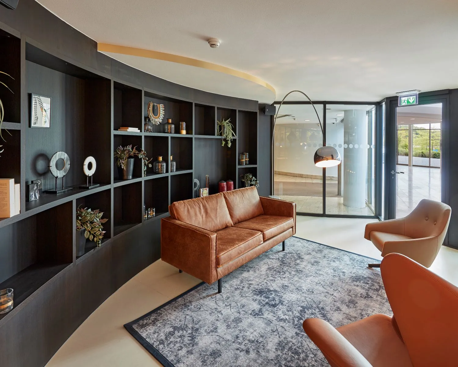 Moderne wachtruimte met een leren bank, designstoelen, wandkast en staande lamp bij glazen ingang.