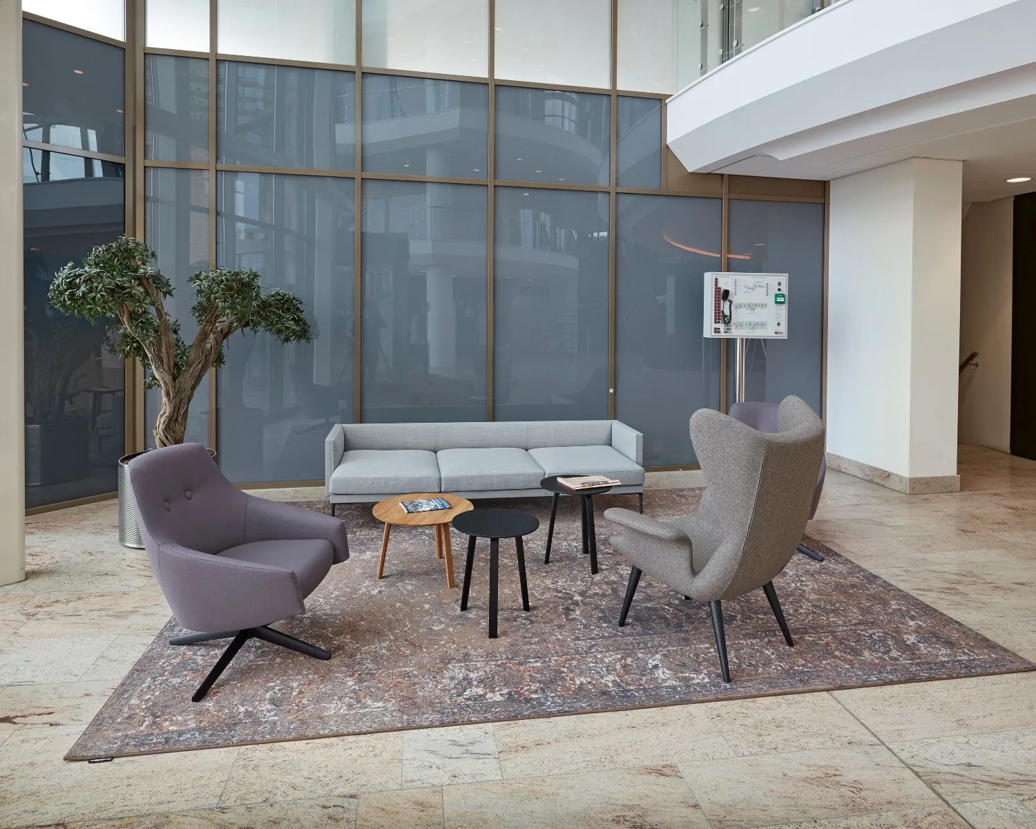 Moderne wachtruimte met grijze zitmeubelen, een vloerkleed en een grote plant in een lichte kantooromgeving.