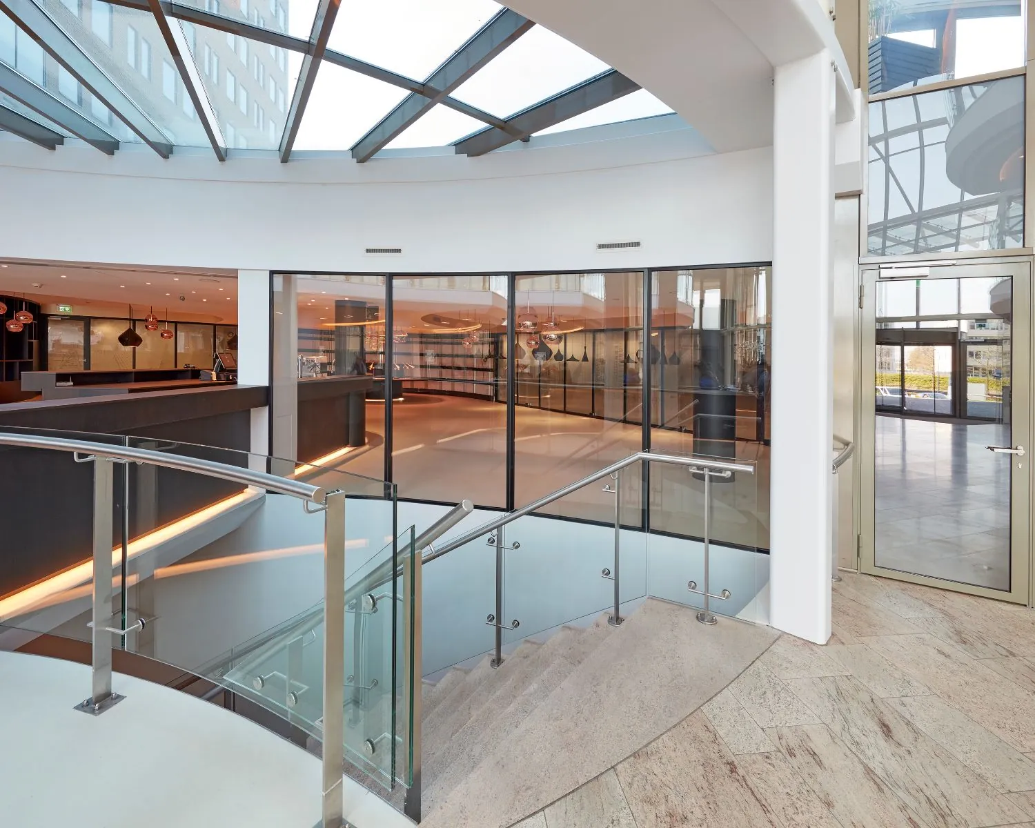 Moderne, lichtovergoten hotellobby met glazen trapleuning en grote ramen.