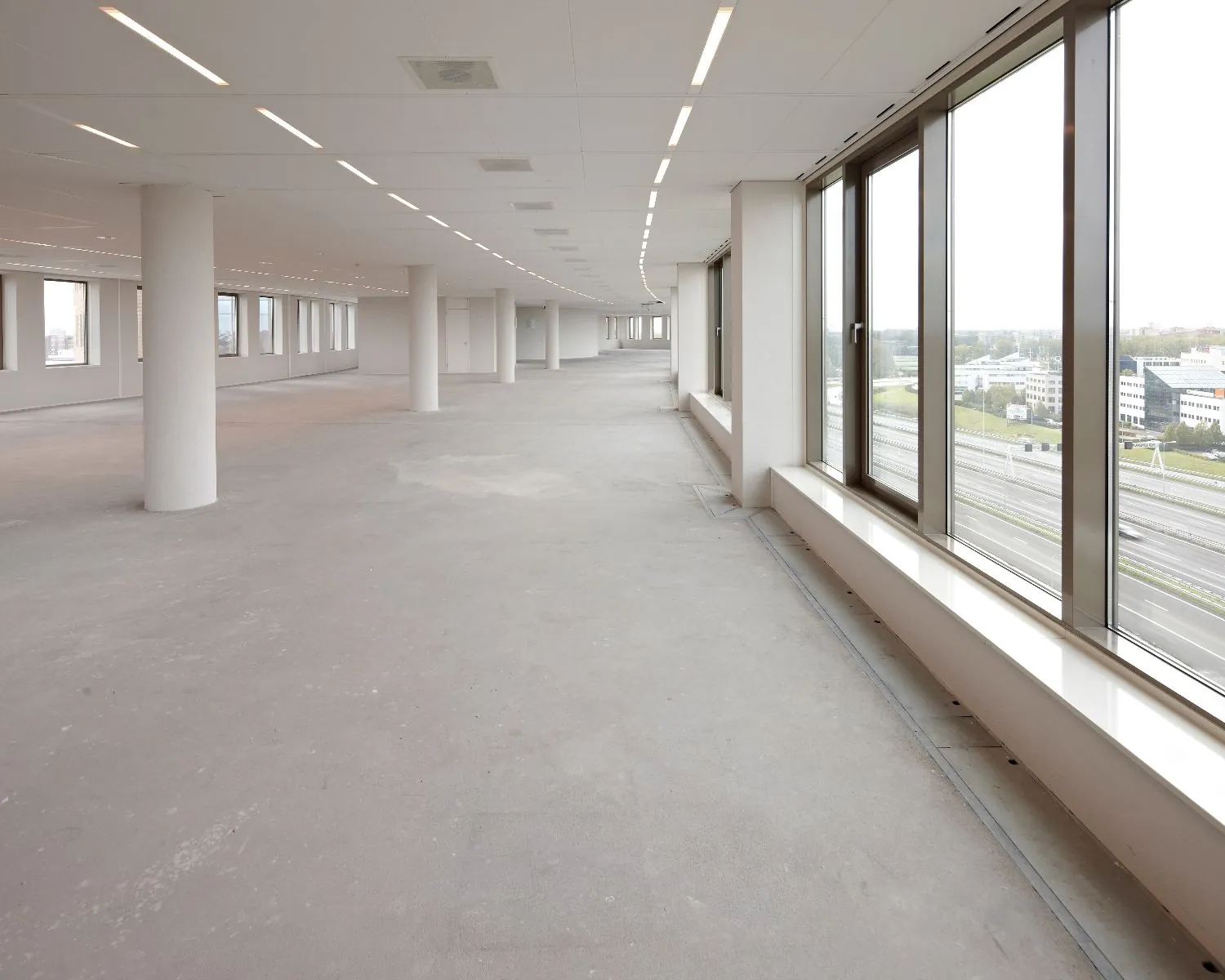 Lege, moderne kantoorruimte met grote ramen en uitzicht op een snelweg en kantoorgebouwen.