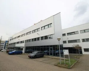 Wit en grijs kantoorgebouw met meerdere verdiepingen en geparkeerde auto's op een parkeerterrein.