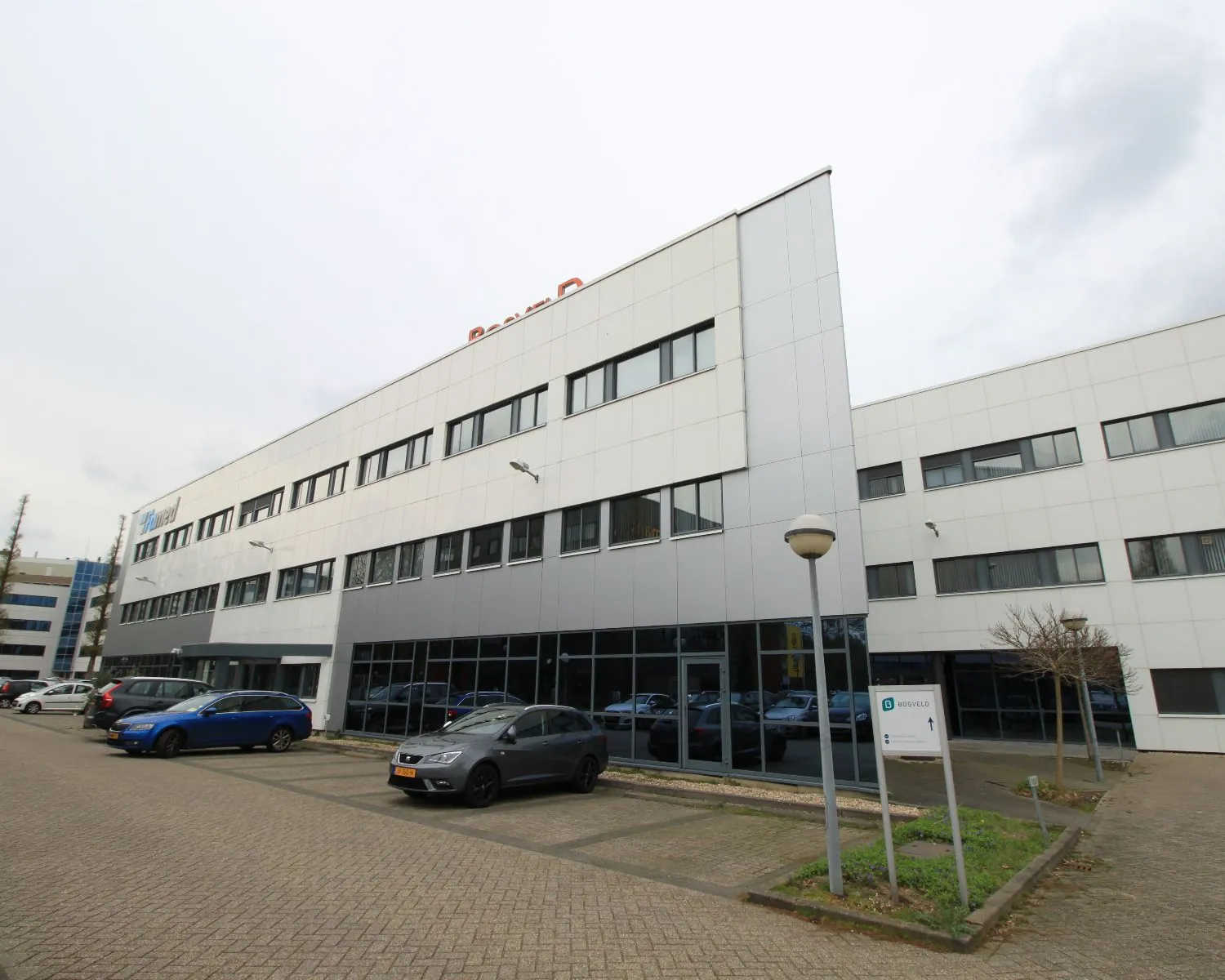 Wit en grijs kantoorgebouw met meerdere verdiepingen en geparkeerde auto's op een parkeerterrein.