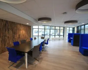 Moderne kantoorruimte met vergadertafel, blauwe stoelen en loungehoeken bij grote ramen.