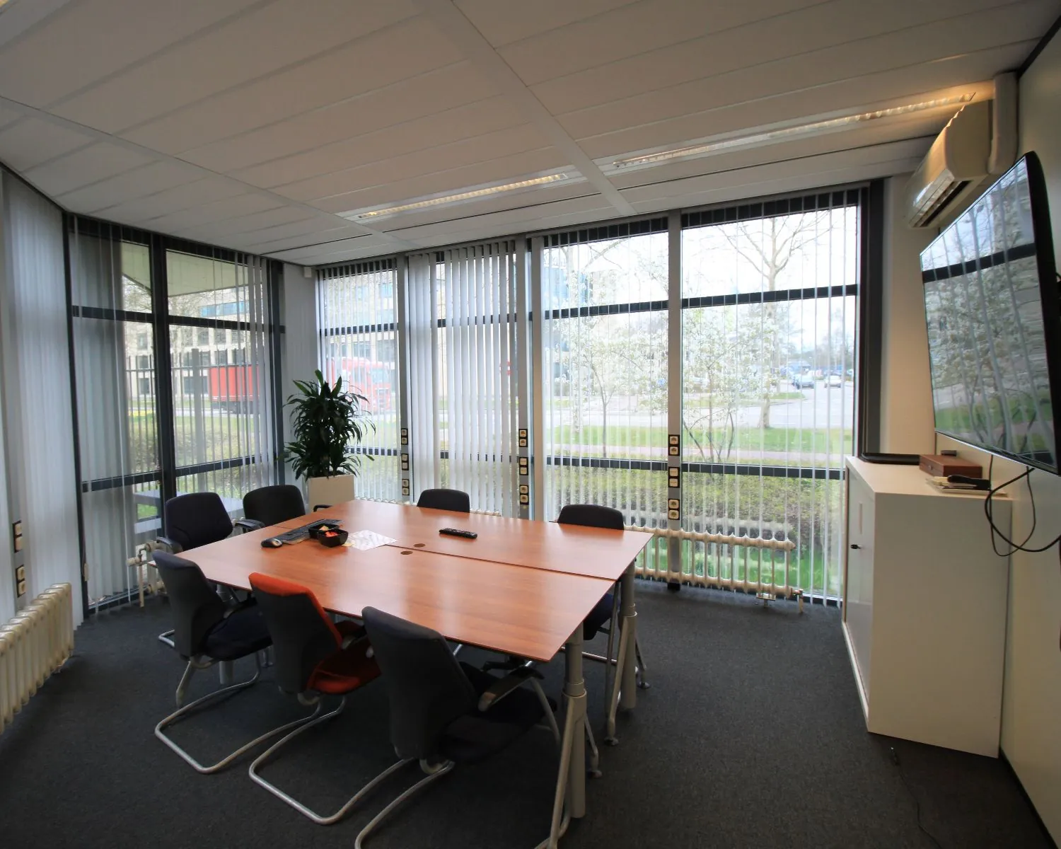 Vergaderruimte met een houten tafel, acht stoelen, een groot scherm aan de muur en ramen met verticale lamellen.