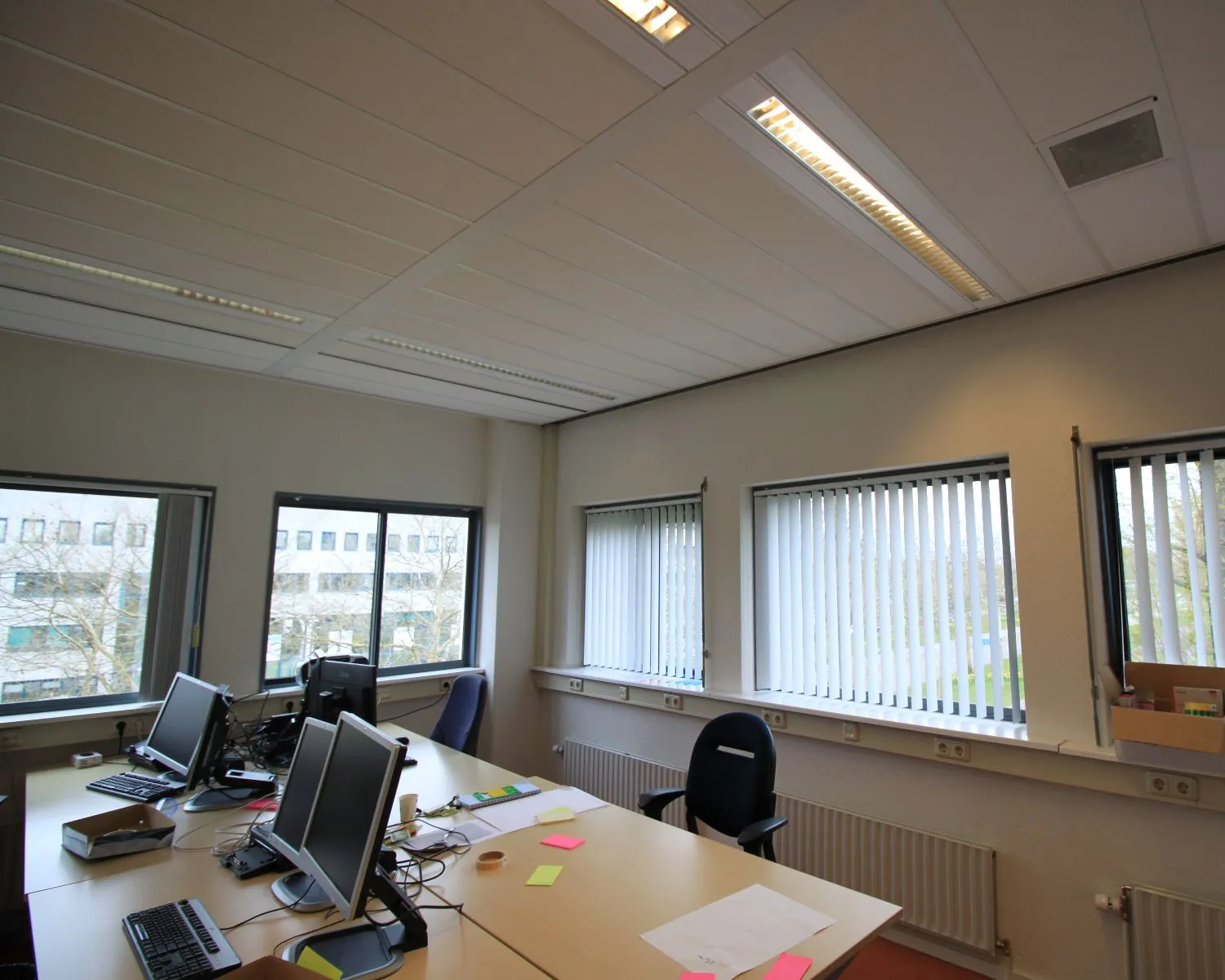 Kantoorruimte met meerdere bureaus, computers en grote ramen met verticale lamellen.